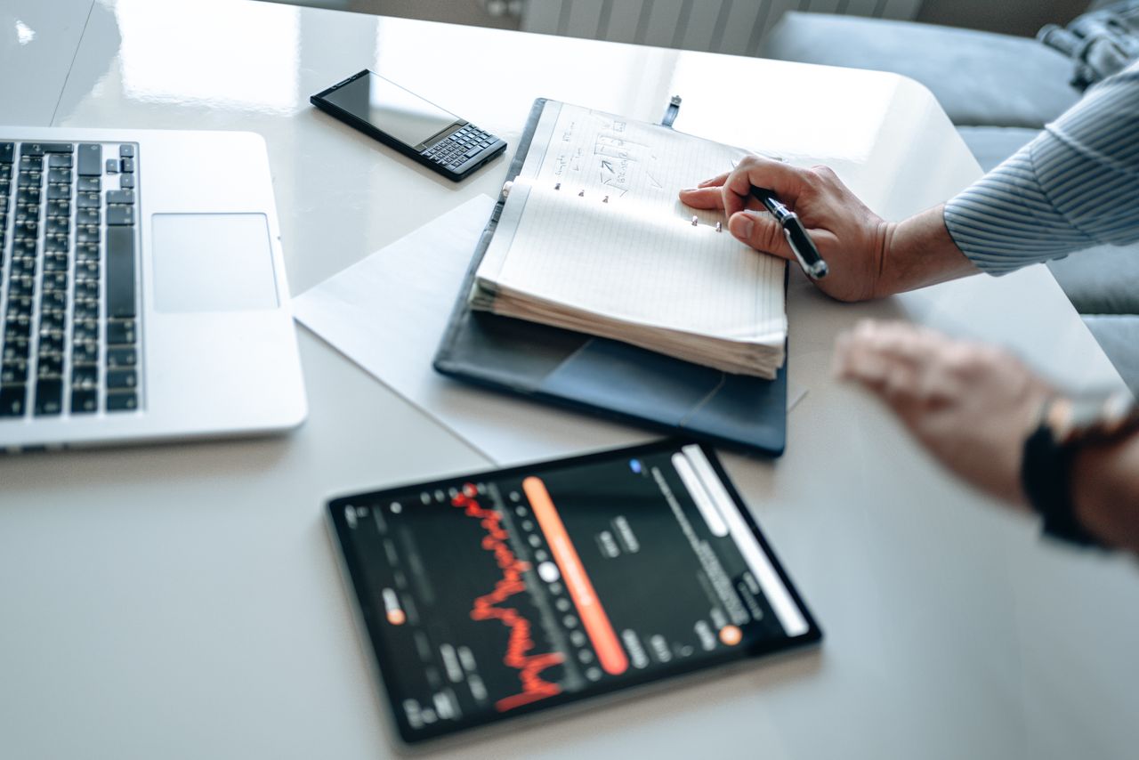 economia da atenção - Uma mesa de trabalho em primeiro plano com alguém tentando digitar ou escrever, mas com os olhos ou a mão inevitavelmente puxados para um smartphone aceso ao lado