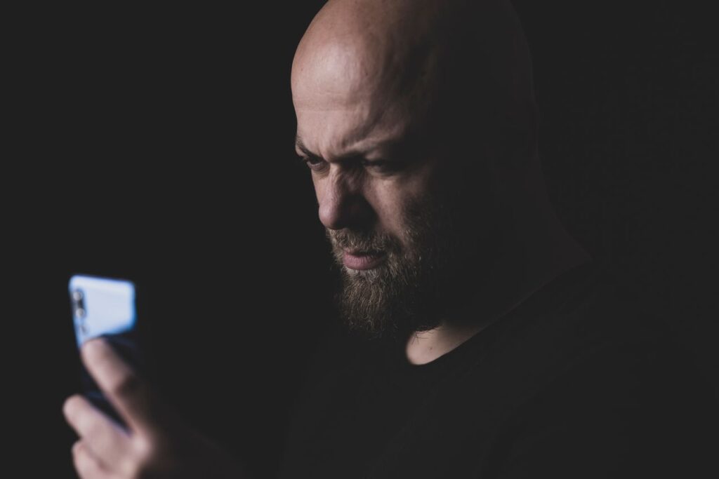 sindrome do pensamento acelerado - Uma pessoa segurando o celular em um ambiente com luz suave ou penumbra, com o rosto levemente iluminado pela tela e uma expressão de cansaço ou sobrecarga