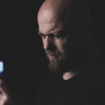 sindrome do pensamento acelerado - Uma pessoa segurando o celular em um ambiente com luz suave ou penumbra, com o rosto levemente iluminado pela tela e uma expressão de cansaço ou sobrecarga