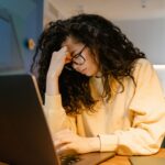 aperfeiçoamento continuo - Uma pessoa visivelmente cansada em uma mesa de trabalho, com a cabeça encostada na mão ou olhando fixamente para a tela de um notebook, cercada de papéis ou xícaras de café