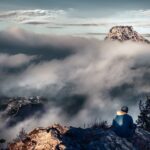 A última das liberdades humanas - Uma pessoa vista de costas ou de perfil, no topo de uma montanha ao amanhecer ou diante de uma paisagem serena e vasta, com iluminação natural suave